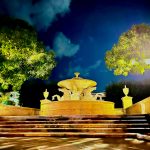 Illuminated ornate fountain at night with dramatic blue sky and trees