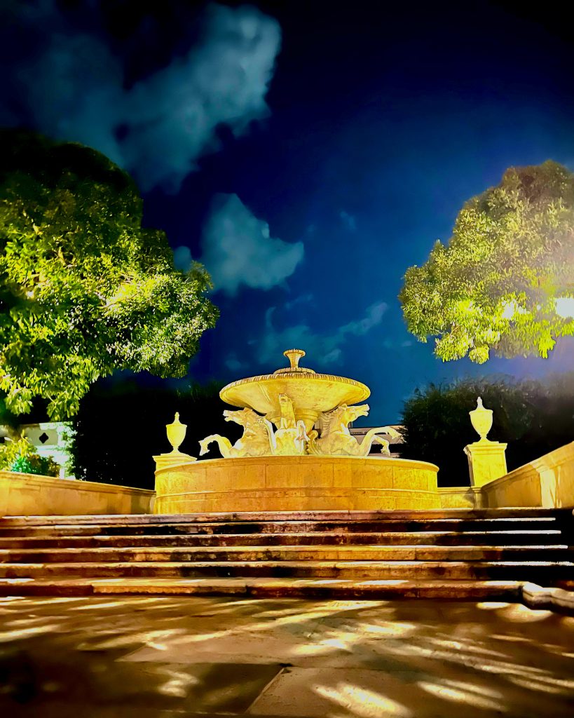 Illuminated ornate fountain at night with dramatic blue sky and trees